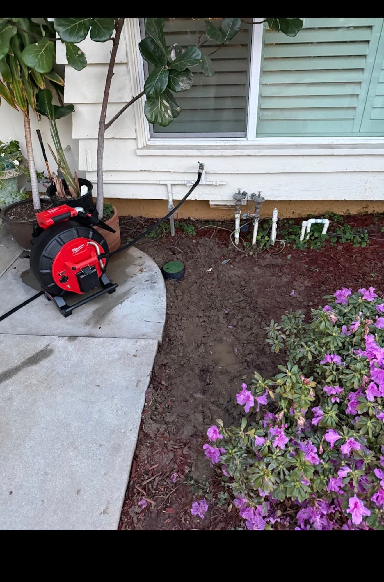Red Milwaukee sewer camera on a concrete path next to muddy ground and house plumbing.