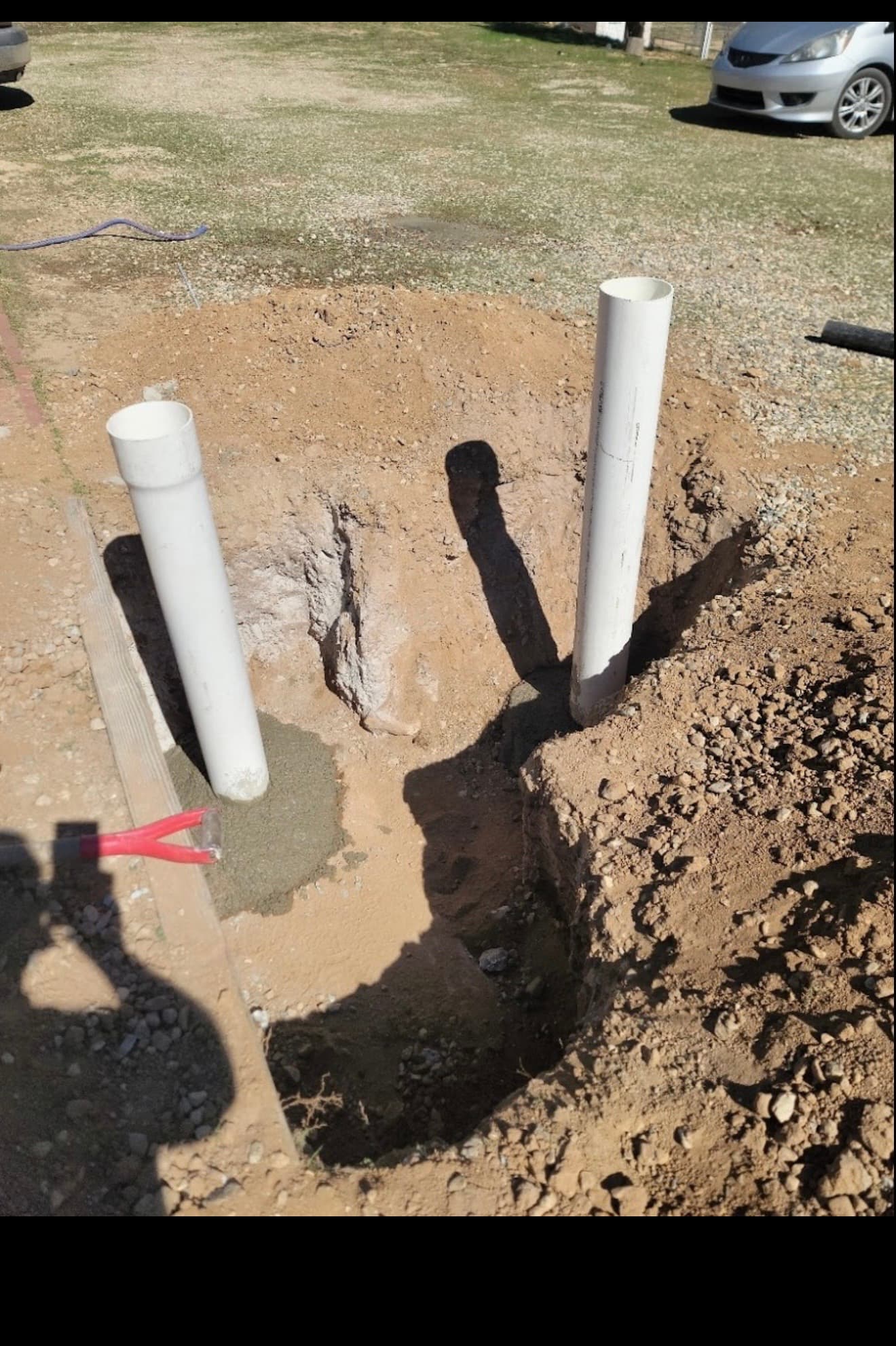 Two white PVC pipes installed in a dirt hole with wet concrete and a shovel.