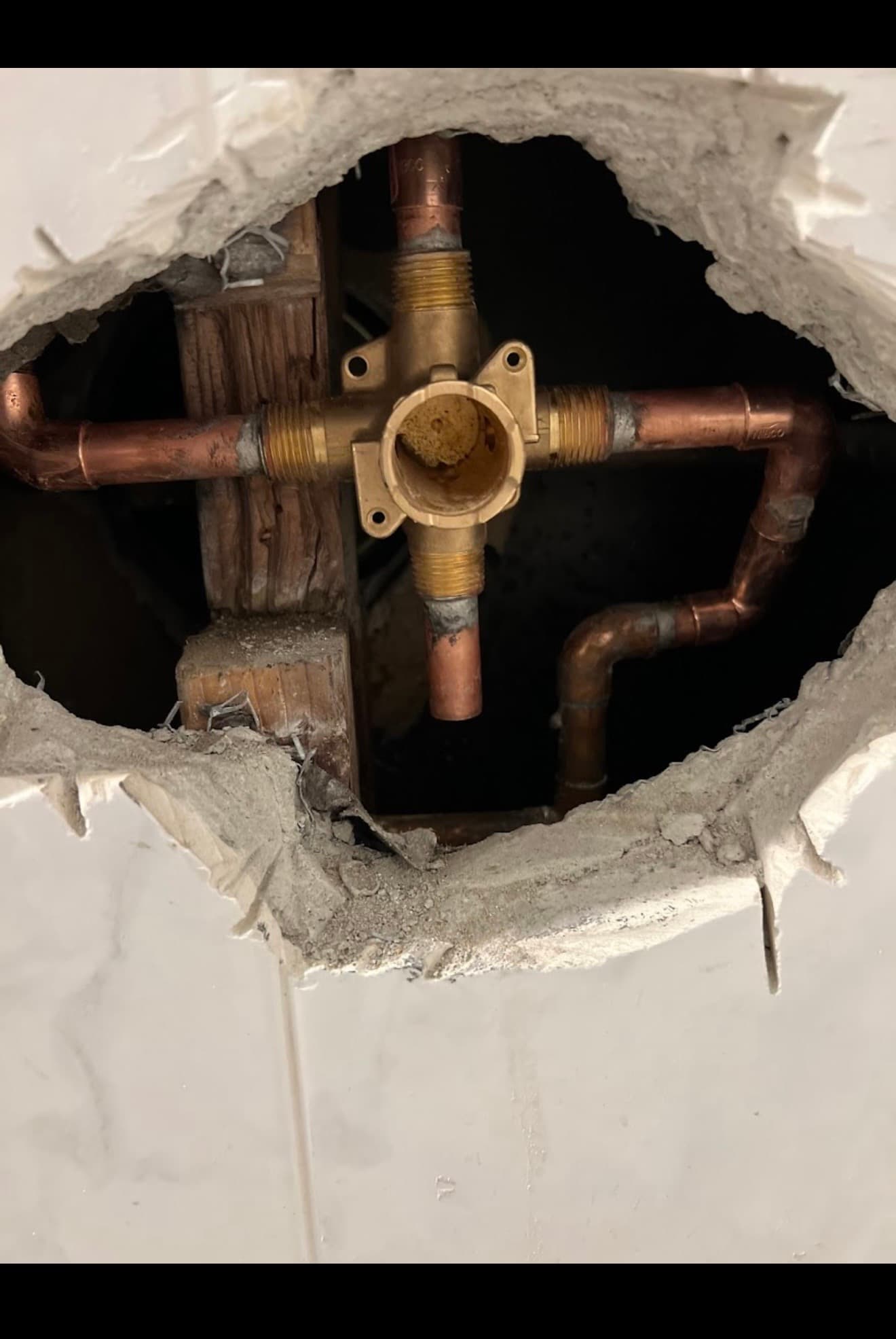 Exposed brass shower valve and copper pipes inside a jagged hole in a tiled wall.