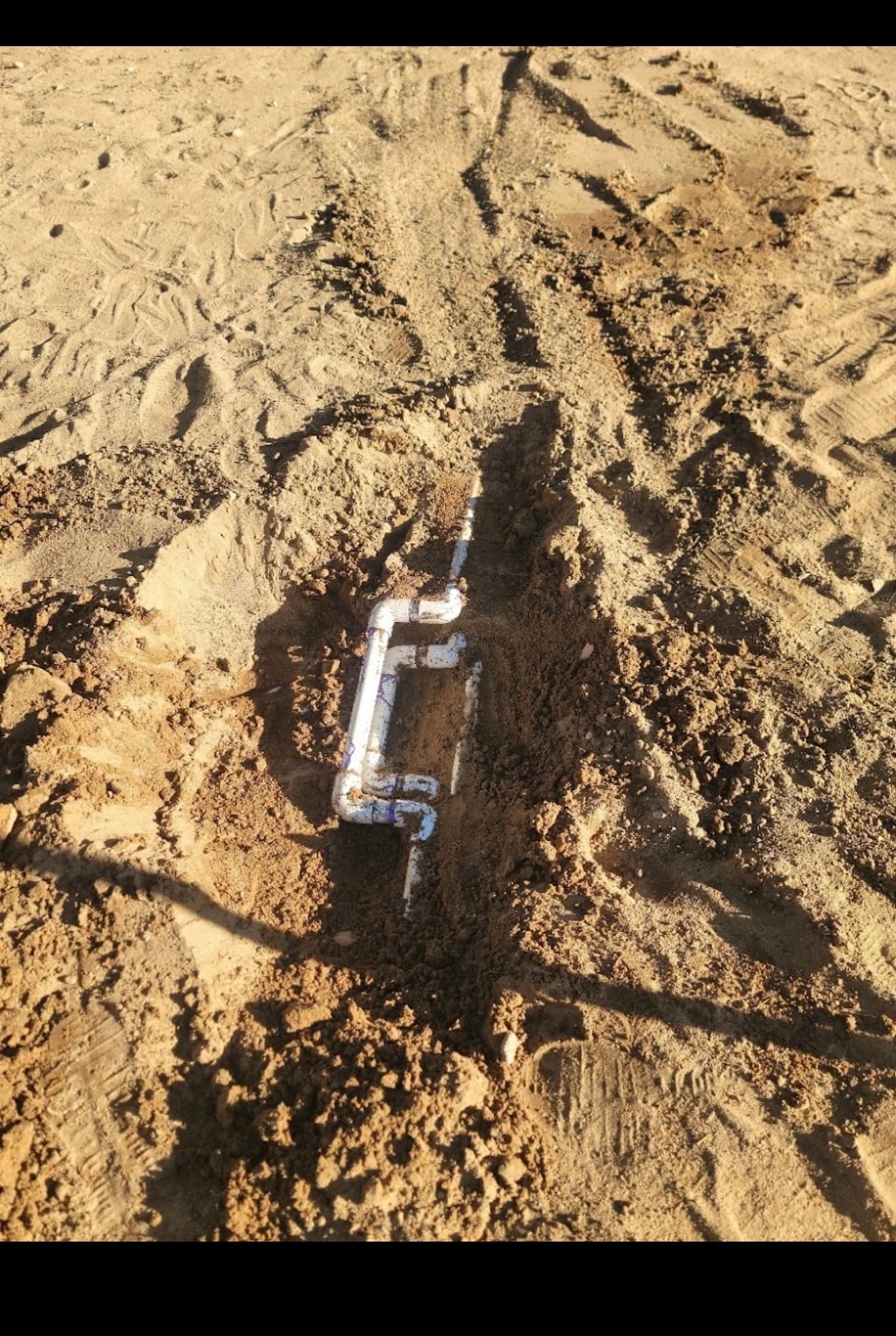 White PVC plumbing pipes partially buried in a rectangular configuration within a sandy dirt trench.