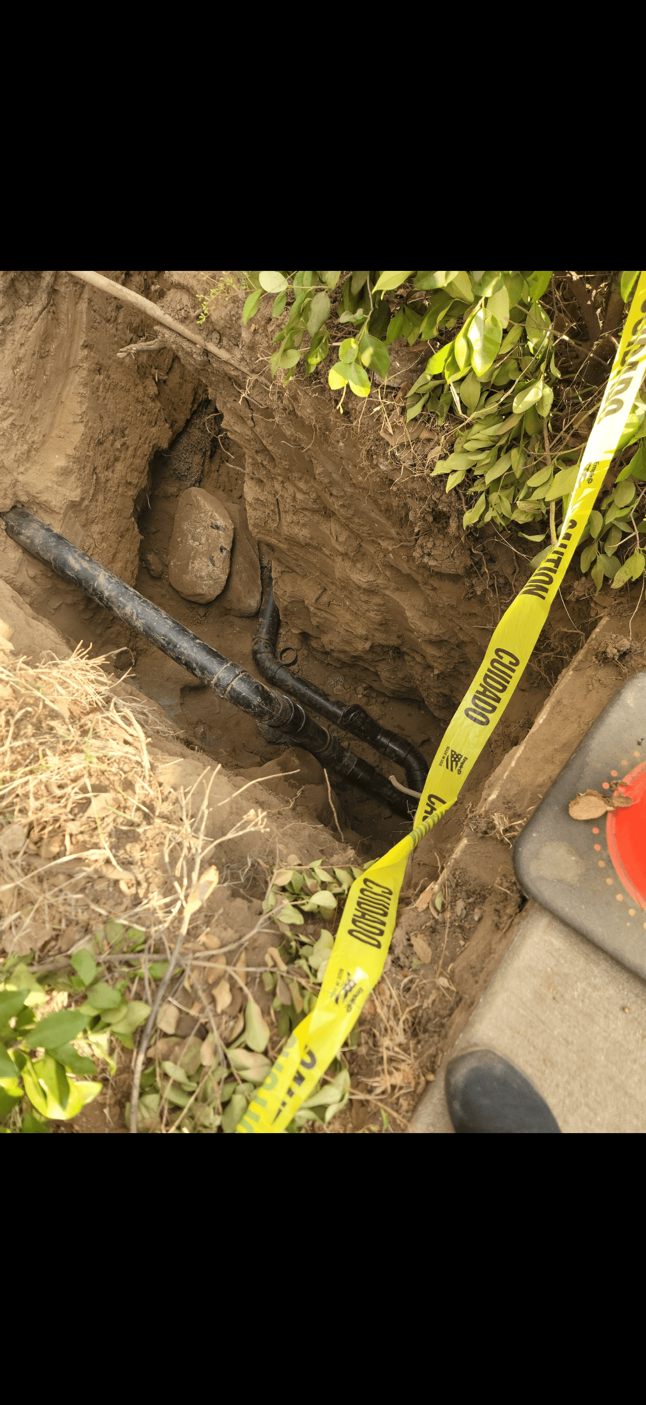 Exposed black pipes in a deep dirt trench marked with yellow caution tape and foliage.