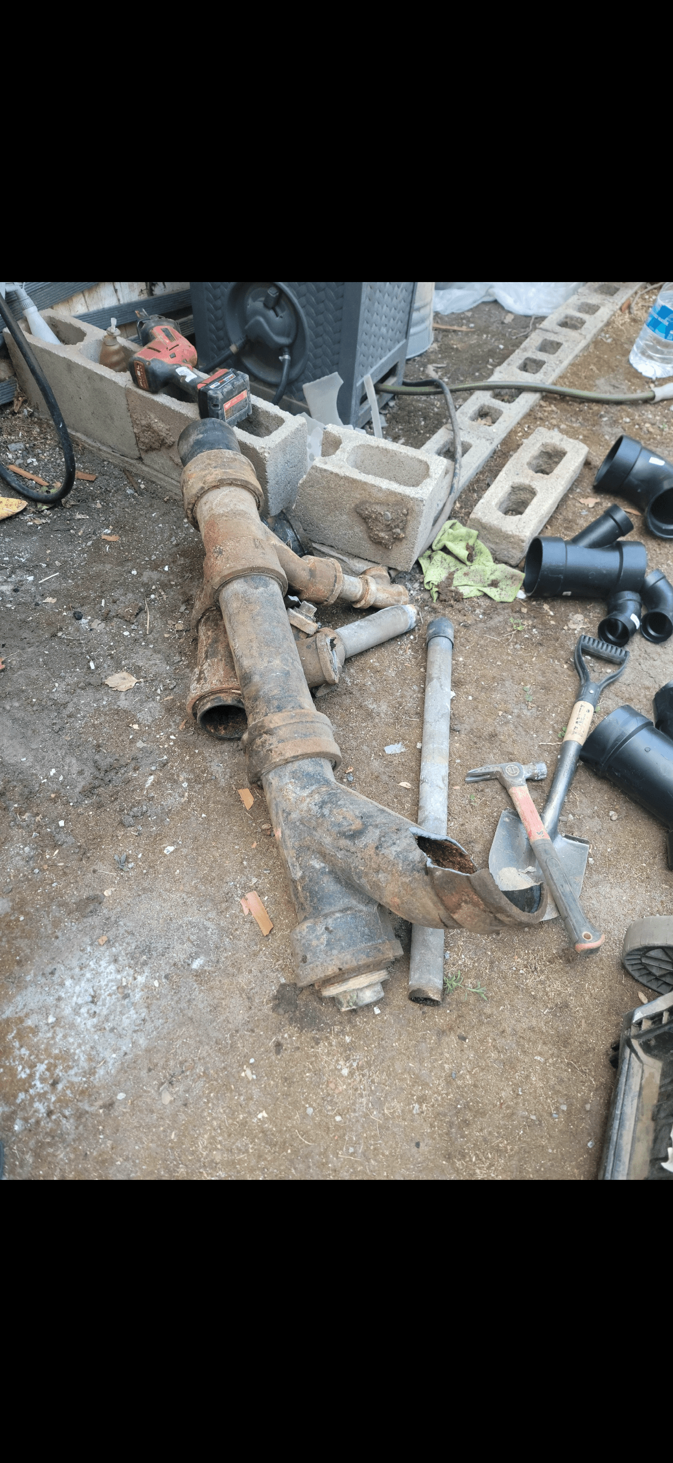 Corroded cast iron pipes and tools lie on dirt near new black PVC fittings.